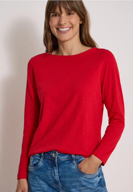 Frau mit langen braunen Haaren in rotem Langarmshirt und Jeans, lächelnd vor neutralem Hintergrund