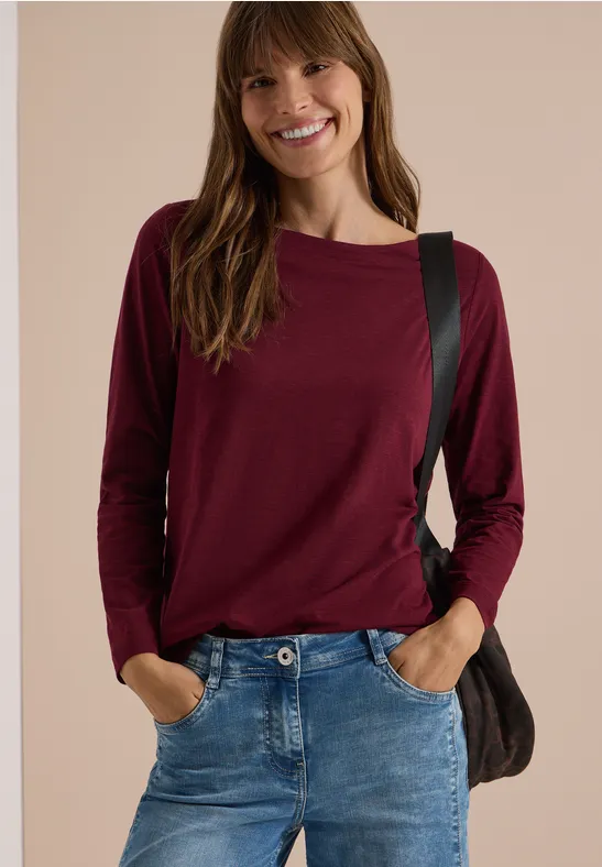 Junge Frau in Jeans und bordeauxrotem Langarmshirt, lächelt, Hände in Hosentaschen, trägt Schultertasche.