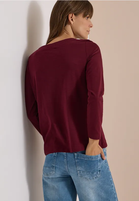 Eine Frau lehnt an einer Wand, trägt einen burgunderfarbenen Langarmpullover und Jeans und steht seitlich zur Kamera.