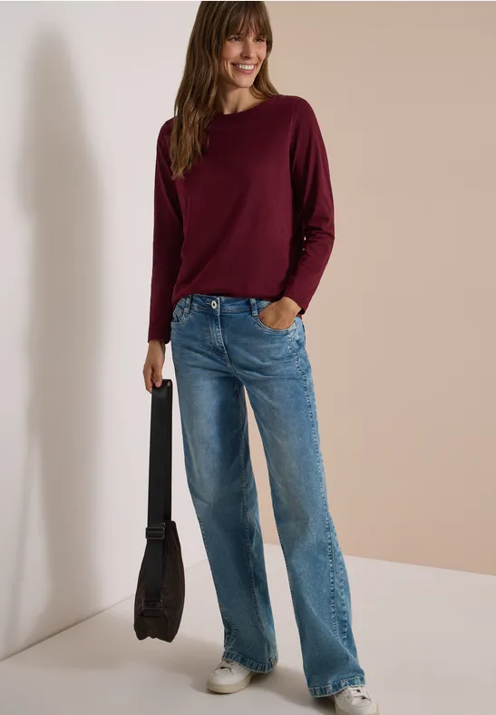 Frau in burgunderrotem Pullover, weitem blauen Jeans und schwarzer Tasche steht vor neutralem Hintergrund.