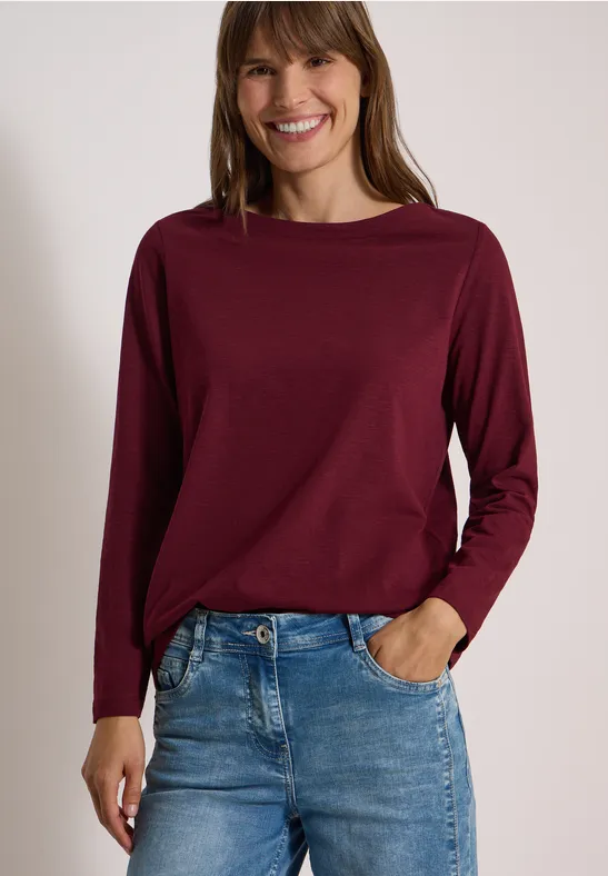 Eine lächelnde Frau trägt ein bordeauxfarbenes Langarmshirt und Jeans.