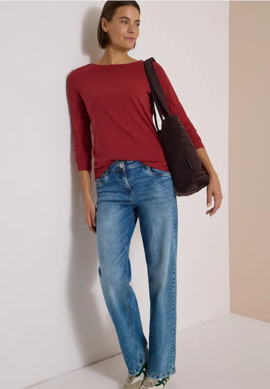 Eine Frau in rotem Langarmshirt, blauen Jeans und Tasche steht lässig an einer Wand.