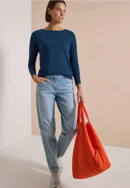 Frau in blauem Langarmshirt, hellen Jeans und weißen Schuhen trägt eine große rote Tasche vor neutralem Hintergrund.