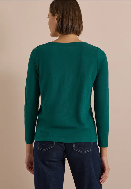 Frau steht von hinten, trägt grünen Pullover und dunkle Jeans vor neutralem Hintergrund.