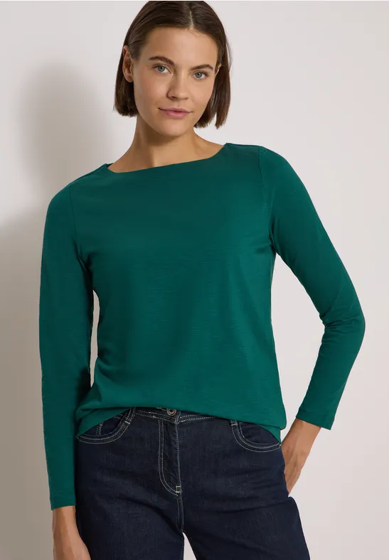 Frau mit grünem Langarmshirt und dunkler Jeans vor hellem Hintergrund.