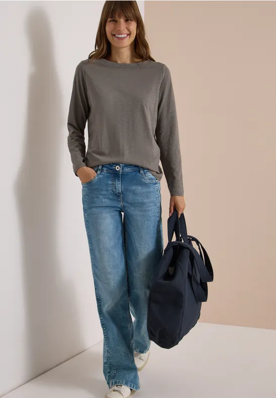 Frau in grauem Langarmshirt, blauen Jeans und weißen Sneakers trägt dunkelblaue Tasche.