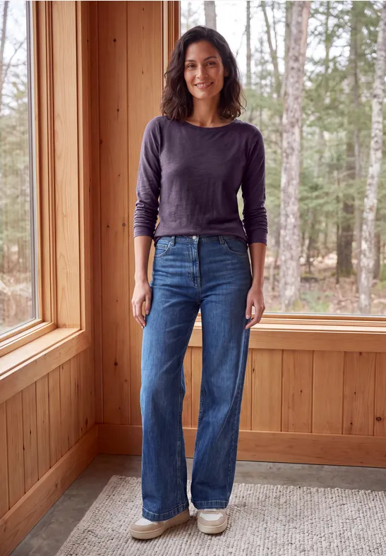Frau steht in einem Holzraum am Fenster, trägt Langarmshirt, weite Jeans und Sneakers.