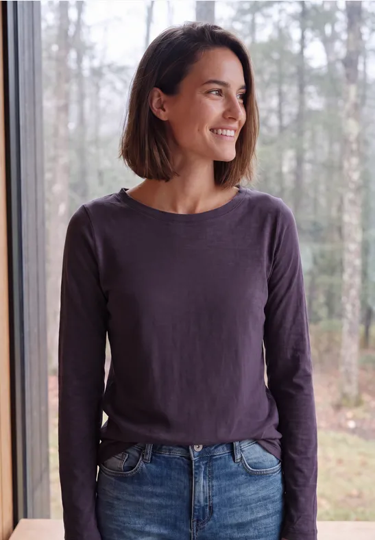 Eine lächelnde Frau mit braunem Bob, lila Longsleeve und Jeans steht am Fenster vor Wald
