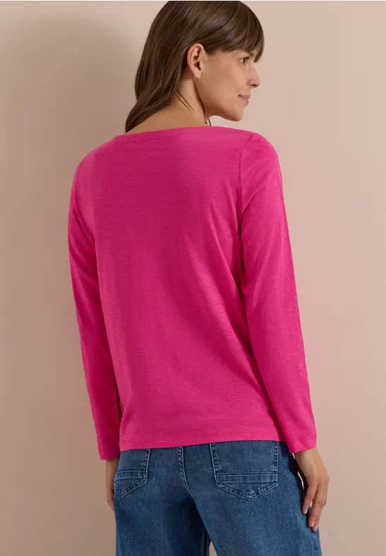 Frau von hinten in pinkem Langarmshirt und blauen Jeans vor beiger Wand.