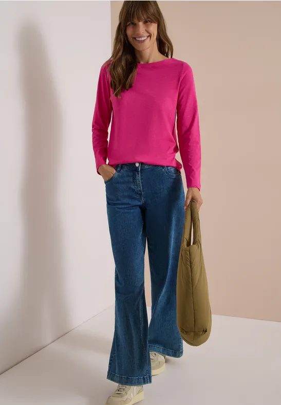 Eine Frau in pinkem Pullover, weitem Jeans und Sneakers lehnt lächelnd an einer Wand und hält eine Tasche.
