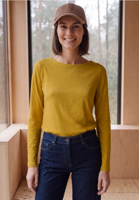 Eine Frau mit braunem Hut, gelbem Langarmshirt und Jeans steht lächelnd vor einem Fenster in einem Holzraum.