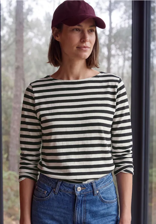 Frau mit roter Baseballmütze, gestreiftem Langarmshirt und Jeans steht vor einem Fenster mit Waldblick.