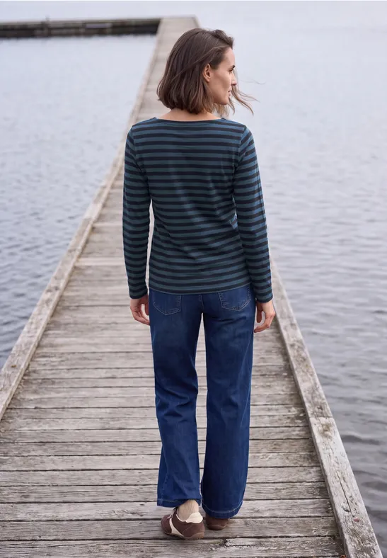 Frau in gestreiftem Pullover und Jeans steht auf einem Holzsteg am Wasser, Blick nach links.