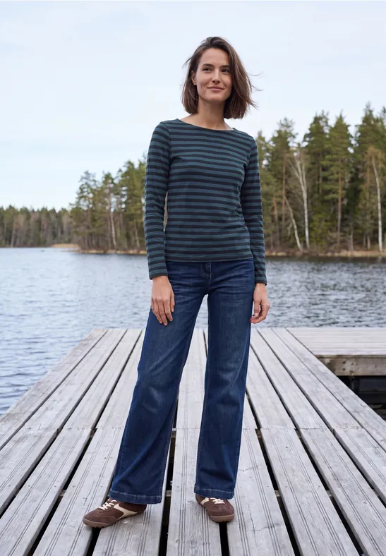 Frau steht auf Holzsteg am See, trägt gestreiftes Shirt und Jeans.