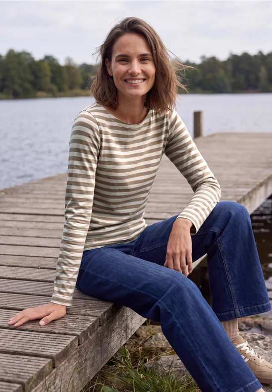 Een lachende vrouw zit op een houten steiger aan een meer, in een gestreept shirt en spijkerbroek.