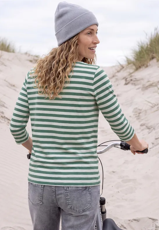 Frau in gestreiftem Shirt und Mütze schiebt Rad über sandige Dünenküste.