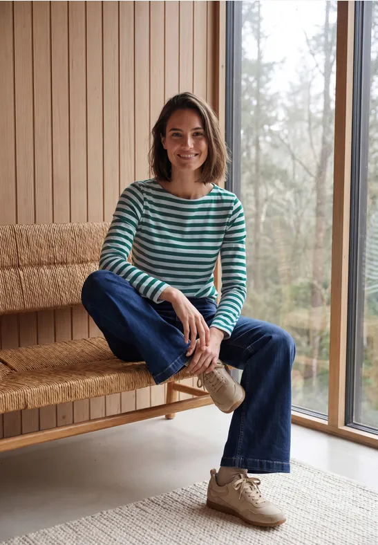 Een lachende vrouw in een gestreepte trui en jeans zit op een houten bank in een lichte kamer.