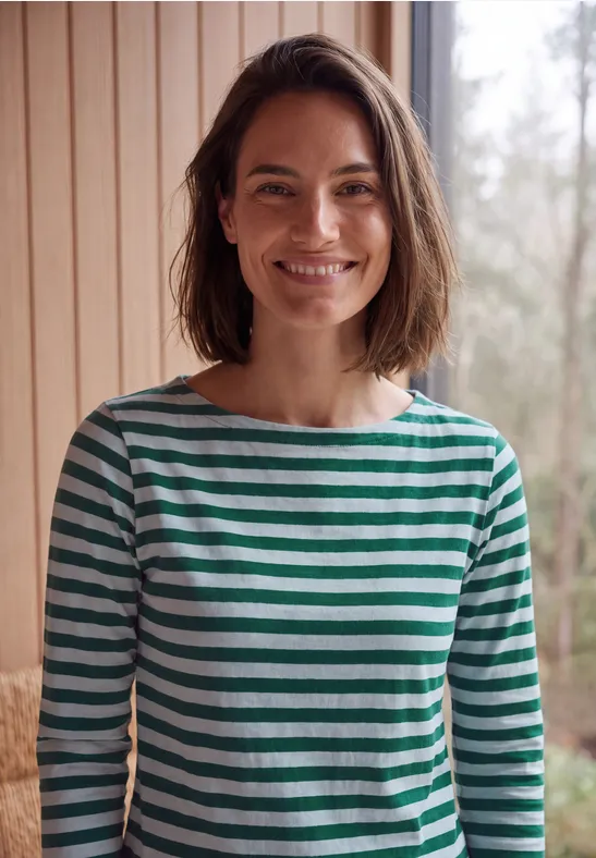 Een vrouw in een groen-wit gestreept shirt lacht voor een licht houten interieur.