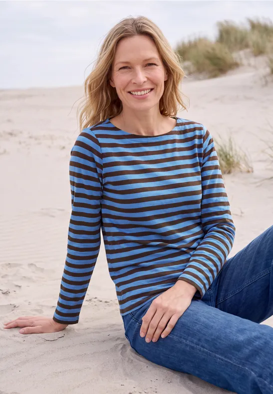 Eine lächelnde Frau sitzt entspannt im Sand am Strand, trägt gestreiften Pullover und Jeans.