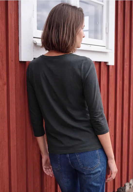 Eine Frau von hinten in schwarzem Oberteil und Jeans lehnt an einer roten Holzwand mit weißem Fenster.