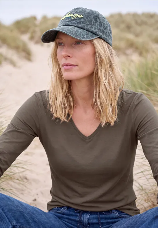 Frau mit Basecap und grünem Shirt sitzt entspannt in einer Dünenlandschaft.