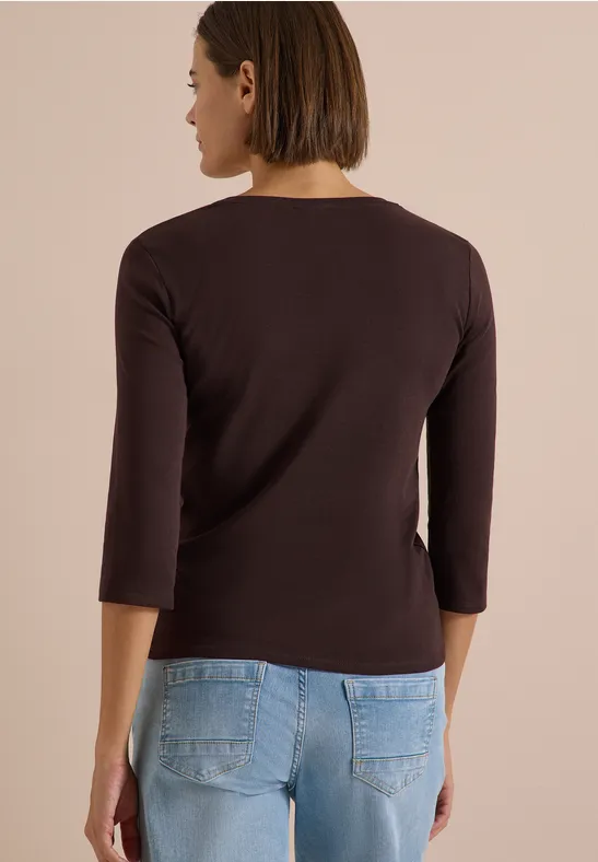 Eine Frau steht mit dem Rücken zur Kamera, trägt ein dunkelbraunes 3/4-Armshirt und blaue Jeans.