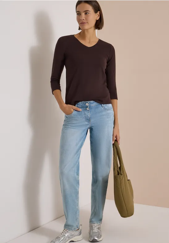 Frau trägt dunkelbraunen V-Ausschnitt-Pullover, helle Jeans, silberne Sneakers und eine olivgrüne Schultertasche.