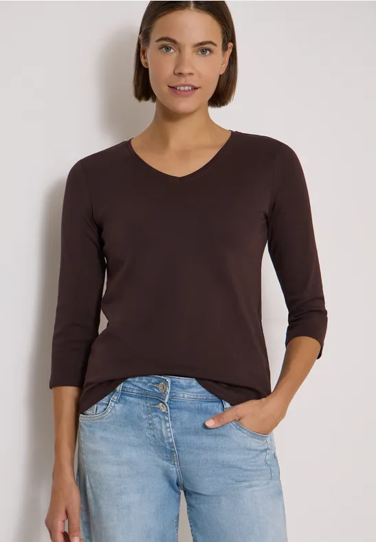 Frau in braunem Langarmshirt und blauen Jeans, Hand in Tasche, lächelnd vor hellem Hintergrund.