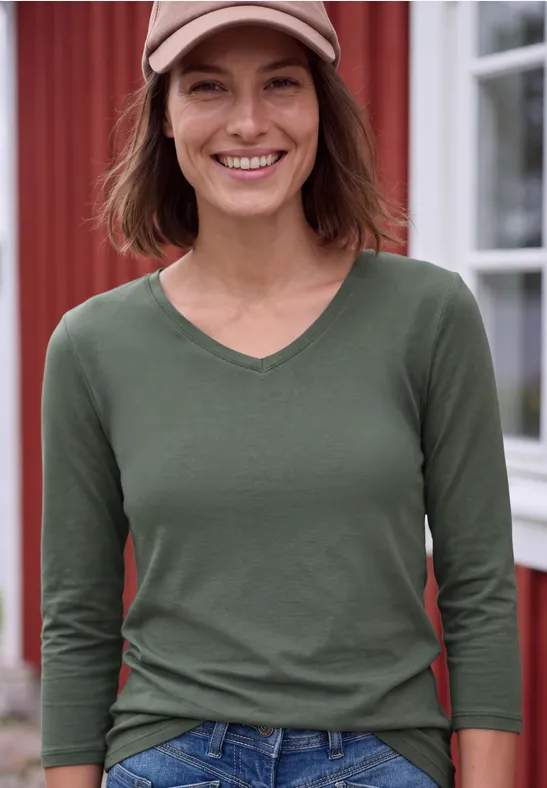 Glimlachende vrouw met groen V-hals shirt en pet, voor een rood gebouw.