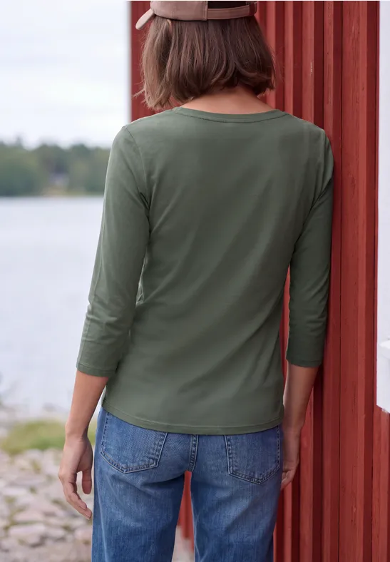 Iemand van achteren met groen 3/4-mouwshirt en spijkerbroek leunend tegen een rood houten muur bij een meer.