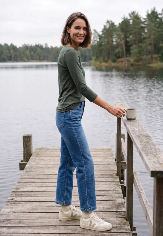 Een glimlachende vrouw in groen shirt en blauwe jeans staat op een steiger bij een meer met een mok.