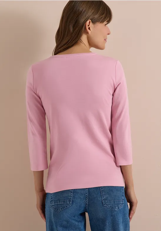 Eine Frau steht mit dem Rücken zur Kamera und trägt ein rosafarbenes 3/4-Arm-Shirt und blaue Jeans.