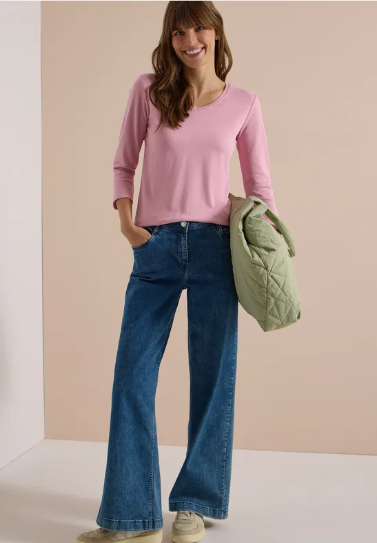 Frau in pinkem V-Pullover, weitem Jeans, weiße Sneaker, hält gesteppte grüne Jacke.