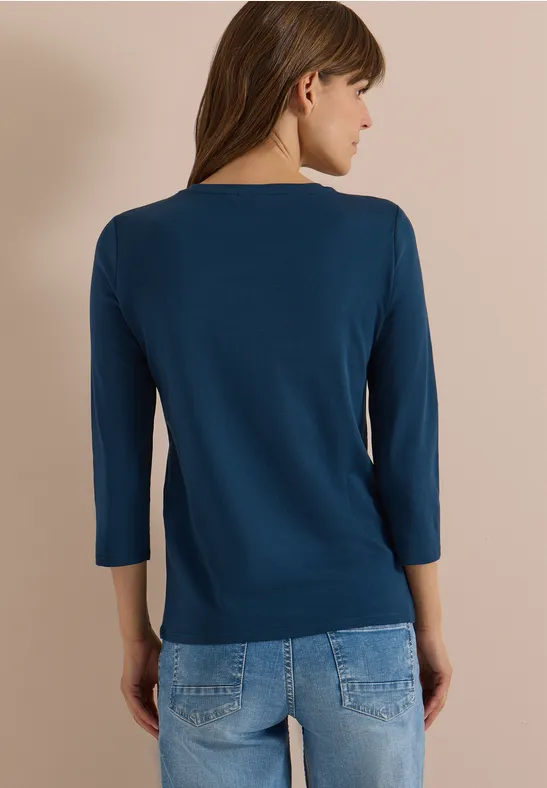 Rückenansicht einer Frau im dunkelblauen 3/4-Arm-Shirt und Jeans vor hellem Hintergrund.