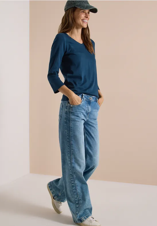 Eine Frau in Jeans, blauem Langarmshirt und Cap, läuft lässig mit Händen in den Taschen.