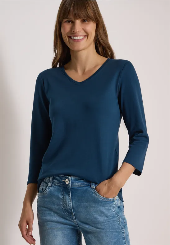 Eine lächelnde Frau mit halblangem Haar trägt ein dunkelblaues V-Shirt und Jeans vor weißem Hintergrund.