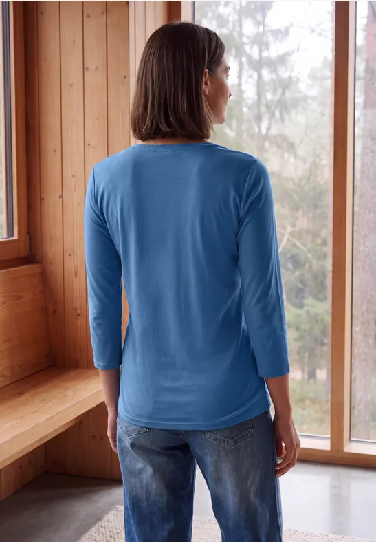 Frau von hinten in blauem Shirt und Jeans, steht in hellem Holzraum vor großen Fenstern mit Blick auf Wald.