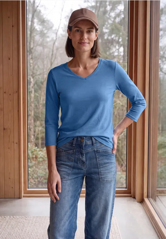 Frau in blauer V-Neck-Strickbluse, Jeans und Cap steht lächelnd vor raumhohen Fenstern aus Holz.