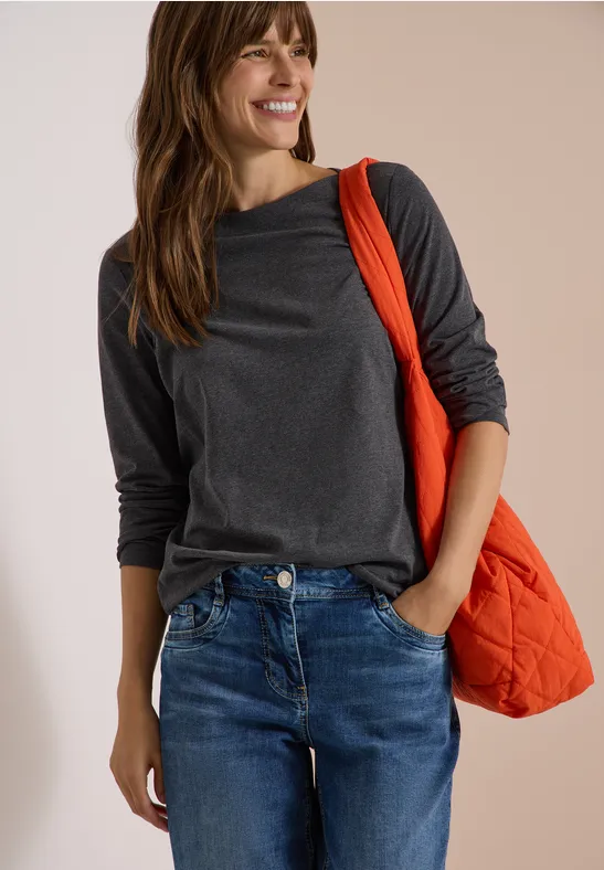 Eine Frau in grauem Langarmshirt und Jeans trägt lächelnd eine orangefarbene Tasche über der Schulter.