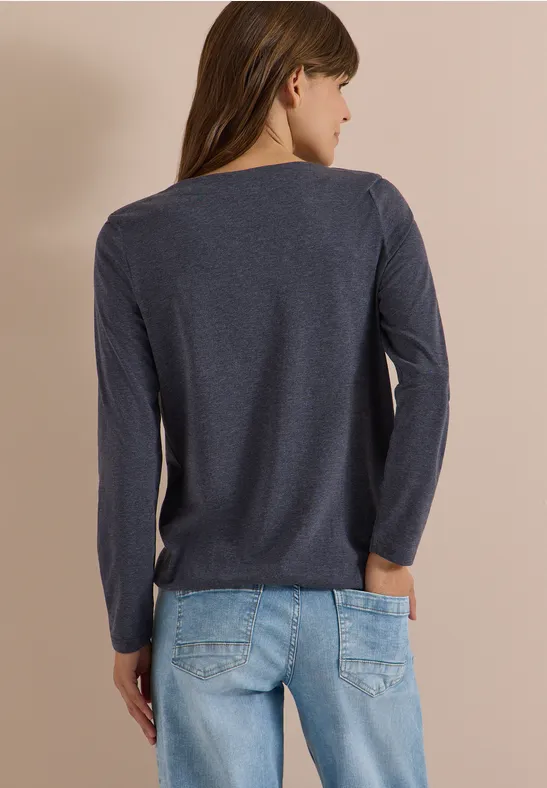 Vrouw van achteren in donkerblauw lang shirt en lichtblauwe spijkerbroek voor een beige achtergrond.