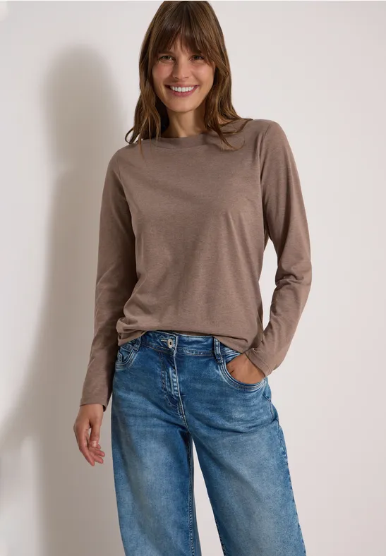 Vrouw glimlacht, draagt een bruin shirt met lange mouwen en blauwe spijkerbroek.