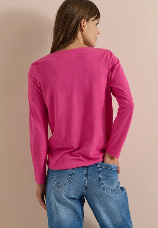 Frau steht von hinten in pinkem Langarmshirt und blauen Jeans vor hellem Hintergrund.