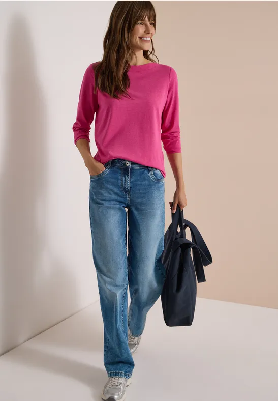Frau mit pinkem Pullover, weiter Jeans und dunkler Tasche steht lächelnd in Zimmerecke.