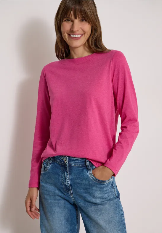 Eine lächelnde Frau in pinkem Langarmshirt und Jeans steht entspannt, eine Hand in der Hosentasche.