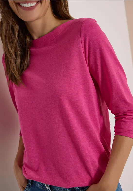 Frau in locker sitzendem pinken 3/4-Arm-Shirt, lächelnd mit Händen in den Taschen.