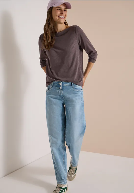 Junge Frau mit Cap, lässigem braunem Shirt, weitem Jeans und Sneakern, lächelnd vor neutralem Hintergrund.
