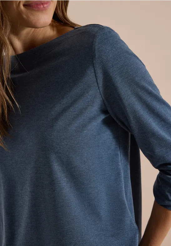 Nahaufnahme einer Frau in blauem Langarmshirt mit weitem Ausschnitt, von Kinn bis Ellenbogen zu sehen.