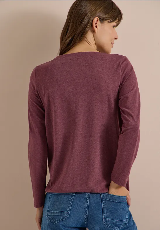 Eine Frau steht seitlich abgewandt im purpurnen Langarmshirt und blauer Jeans vor beigem Hintergrund.