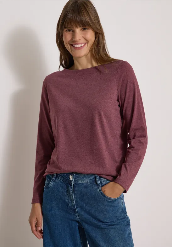 Lächelnde Frau in bordeauxrotem Langarmshirt und blauen Jeans vor hellem Hintergrund.
