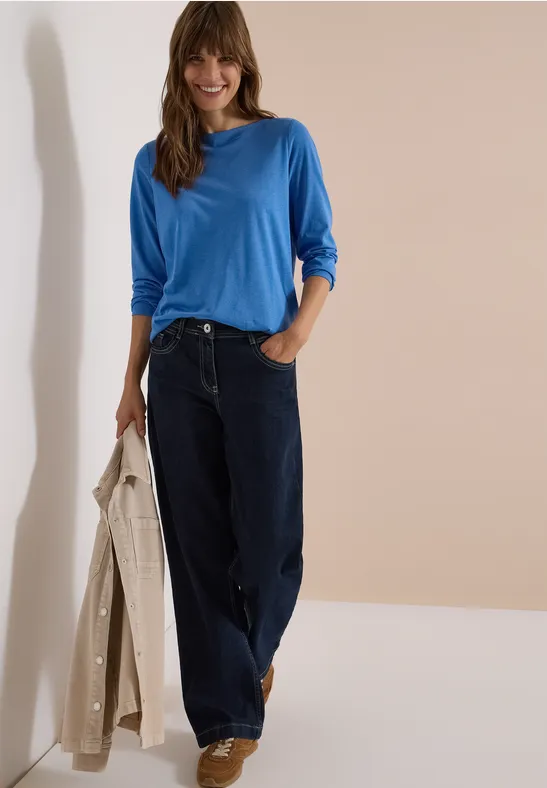 Frau in blauem Langarmshirt und weiten Jeans, hält beigen Mantel, steht vor hellem, minimalistischen Hintergrund.